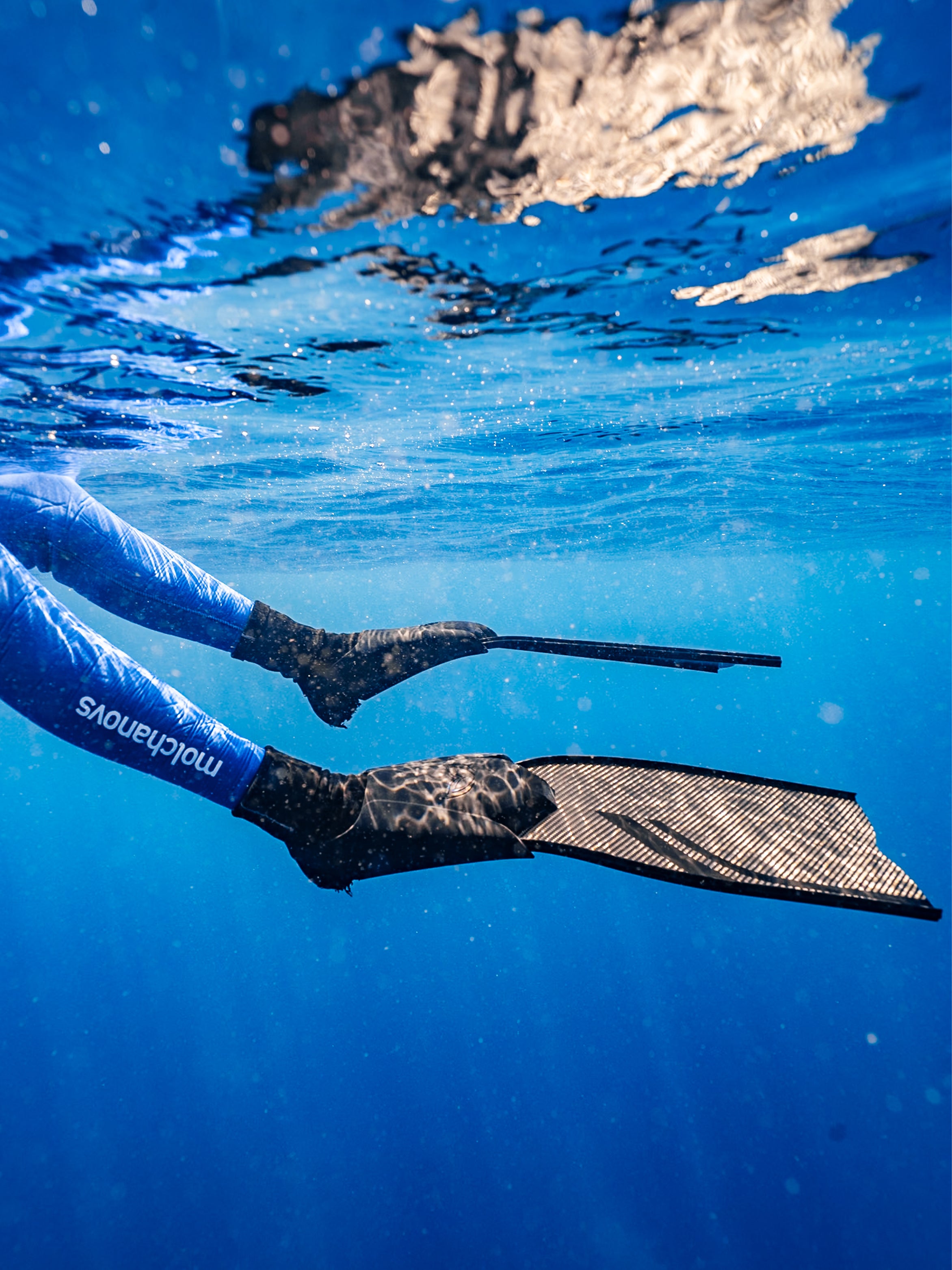 Freediver wearing Molchanovs wetsuit kicking with short carbon blade fins near the water surface, underwater side view